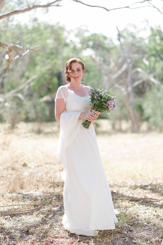 Red headed woman in a lace wedding gown. She is standing in the bush holding a bouquet of flowers.