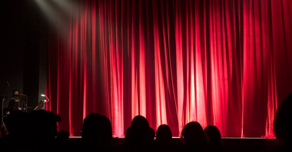 Dimly lit theater stage with red curtains and audience silhouettes under spotlights.